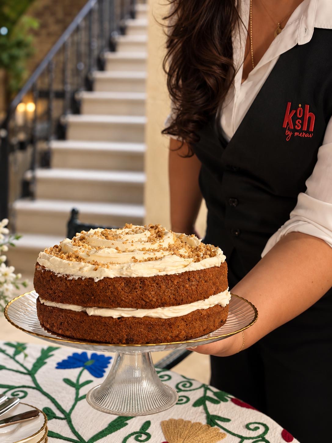Merav in her KOSH uniform presenting a double-layer cream-topped cake on a glass stand in front of a stone staircase.