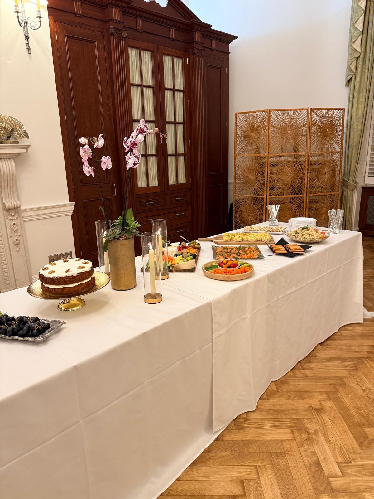 A long white-clothed buffet in a panelled room with a walnut-topped cake on a gold stand, orchids, candles, boards of cheese, salmon and savoury bites.